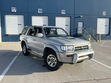 Load image into Gallery viewer, Warm Grey JDM KZN185 Hilux Surf SUV for sale parked in front of a building with numbered garage doors.
