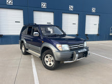 Load image into Gallery viewer, Blue JDM KZJ95 Land Cruiser Prado SUV for sale parked in front of a building with numbered garage doors.
